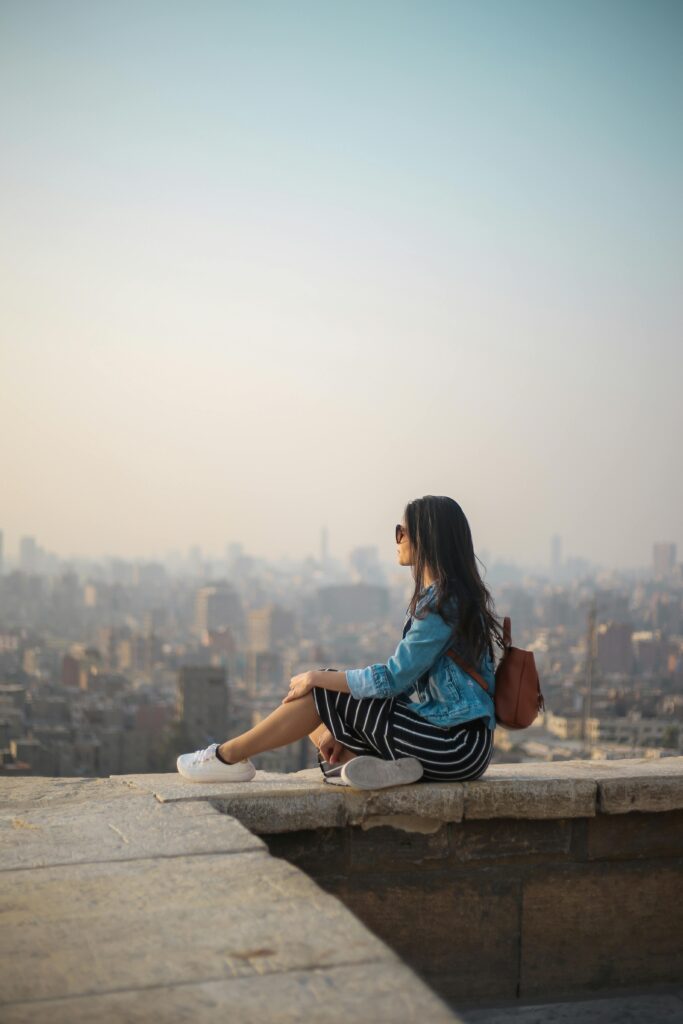 A young woman in casual attire sits on a rooftop terrace, viewing a sprawling cityscape in daylight.